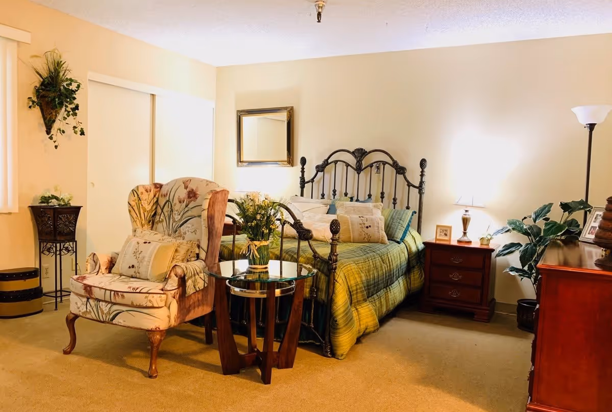 A cozy bedroom with a metal framed bed covered in a green plaid comforter and multiple pillows. Next to the bed is a wooden nightstand with a lamp, a small plant, and framed photos. A floral upholstered armchair with a matching pillow sits beside a round glass-top side table with a flower vase. The room has beige walls, a carpeted floor, a standing lamp, a wall mirror, and several potted plants.