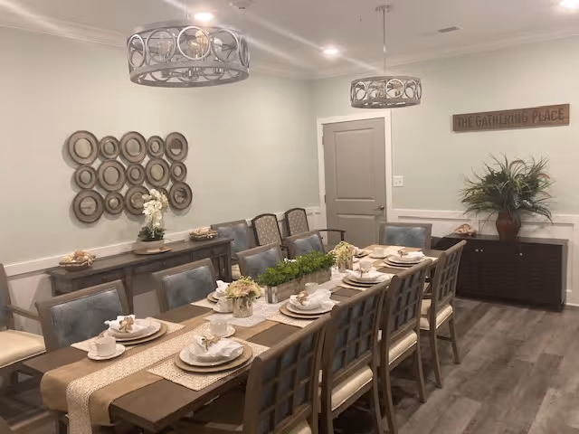 A dining room with a long wooden table set for a meal with plates, cups, and napkins. The table has a beige runner and floral centerpieces. There are multiple chairs around the table. The room has light-colored walls, two modern circular chandeliers, a sideboard with decorative plates on the wall above it, and a cabinet with a plant and a sign that reads 'THE GATHERING PLACE'.