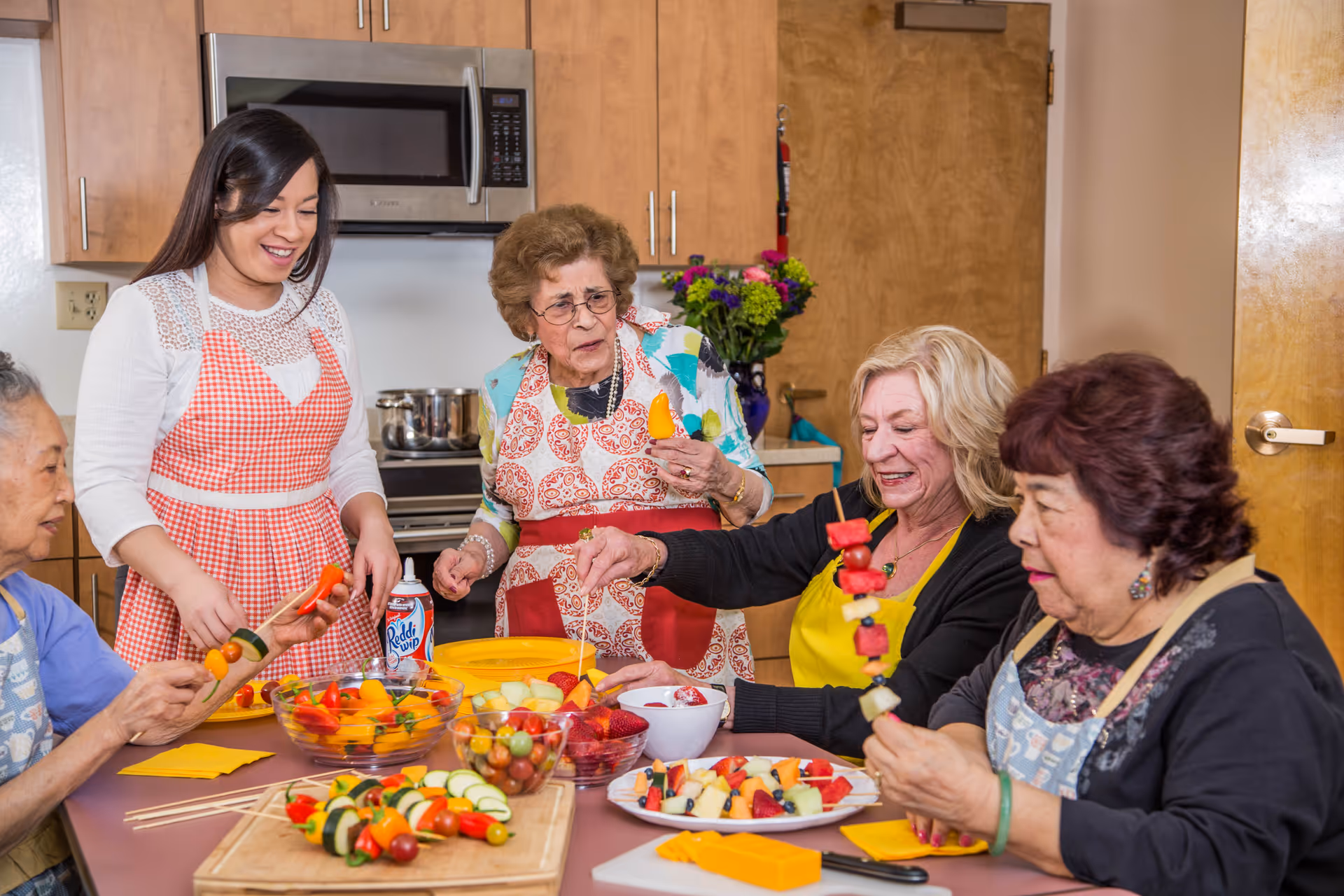 A group of five women, including elderly women and a younger woman, are gathered around a kitchen table preparing colorful fruit and vegetable skewers. They are smiling and interacting with each other in a warm, well-lit kitchen with wooden cabinets and a microwave in the background.