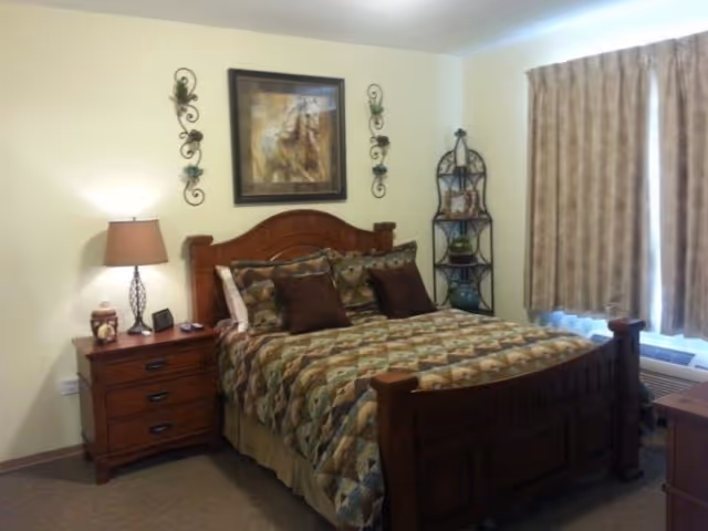 A bedroom with a wooden bed frame and a patterned quilt. There are four pillows on the bed, two of which are dark brown. To the left of the bed is a wooden nightstand with a lamp, a small decorative item, and a digital clock. Above the bed hangs a framed picture of a horse, flanked by two decorative wall sconces. To the right of the bed is a metal corner shelf with decorative items and a window with beige curtains partially drawn. Below the window is an air conditioning unit.