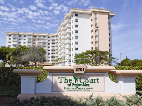 Exterior view of a multi-story senior living facility building behind a sign that reads '2701 The Court at Palm Aire Five Star Senior Living Assisted Living Facility #5611' with trees and a partly cloudy sky in the background.
