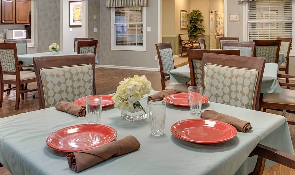A dining area in a senior living facility with tables covered in light blue tablecloths, each set with red plates, brown napkins, and clear drinking glasses. The chairs have wooden frames and patterned upholstery. A small vase with white flowers is placed in the center of the table. The room has wooden flooring and neutral-colored walls with windows and framed artwork.