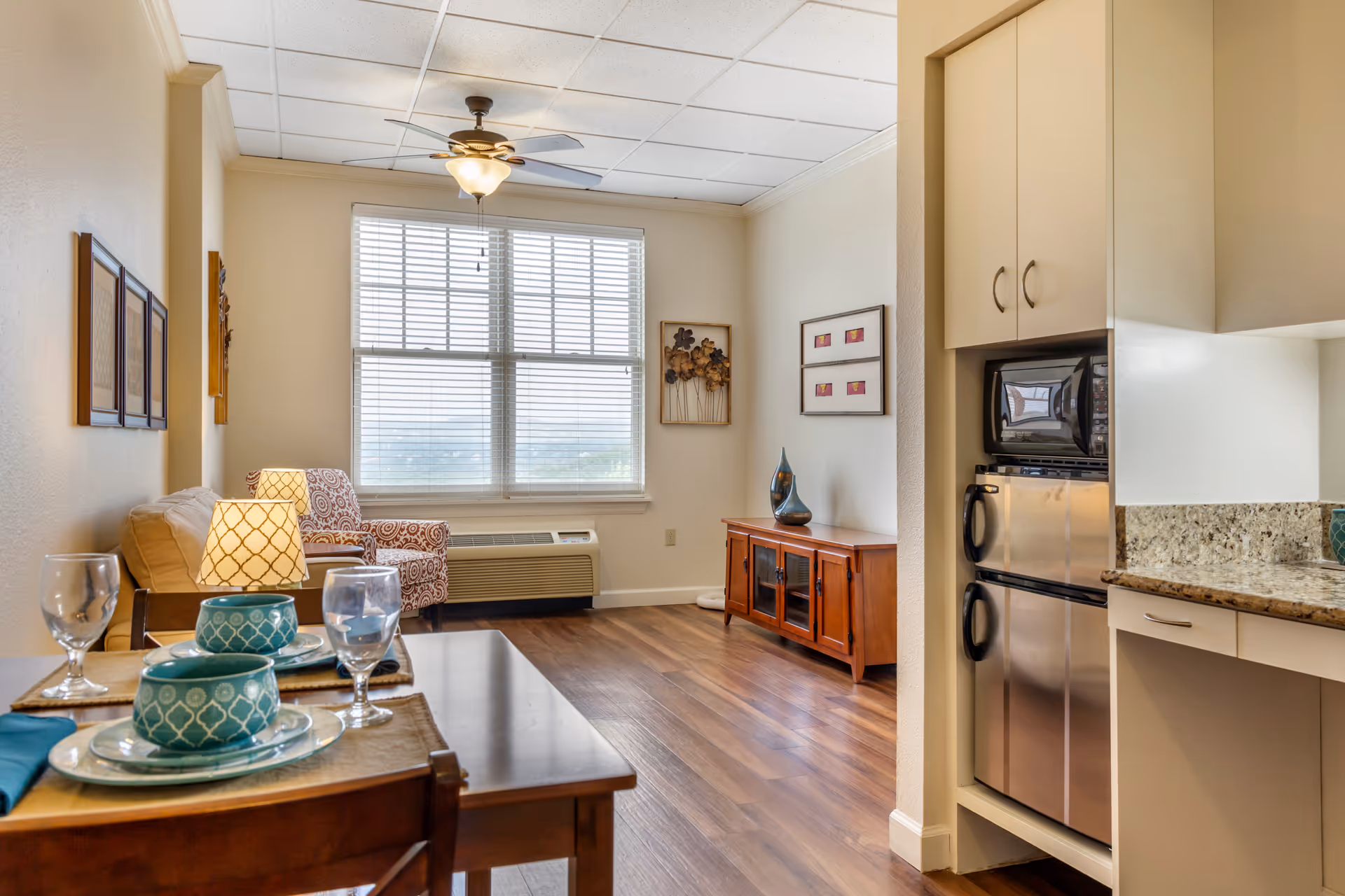Bright living room and kitchenette in a senior living apartment with a set dining table, armchair, TV console, and large window.