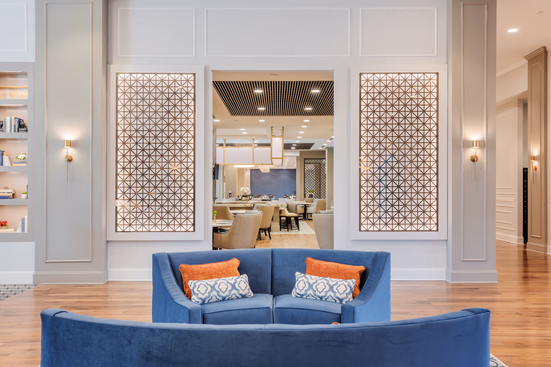 Curved blue sofas with patterned and orange throw pillows face decorative geometric wall panels opening into a dining area in a bright communal lounge.
