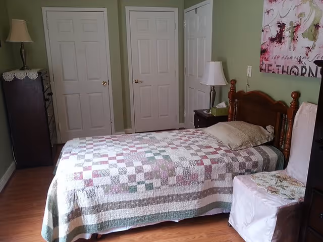 A bedroom with a single wooden bed covered with a patchwork quilt in shades of green, pink, and white. There is a pillow on the bed and a nightstand with a lamp and tissues next to it. The walls are painted light green, and there are three white doors on the far wall. A dresser with a lamp and lace doily is on the left side, and a chair with a white cover is on the right side. A colorful abstract painting hangs on the wall above the bed.