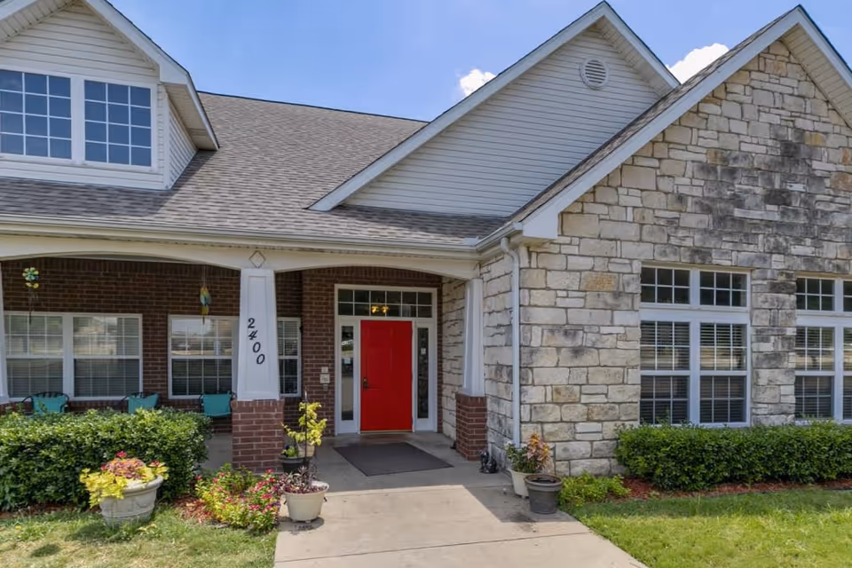 Front exterior view of a senior living facility with a red door entrance, stone and brick facade, large windows, and a covered porch with chairs and potted plants.