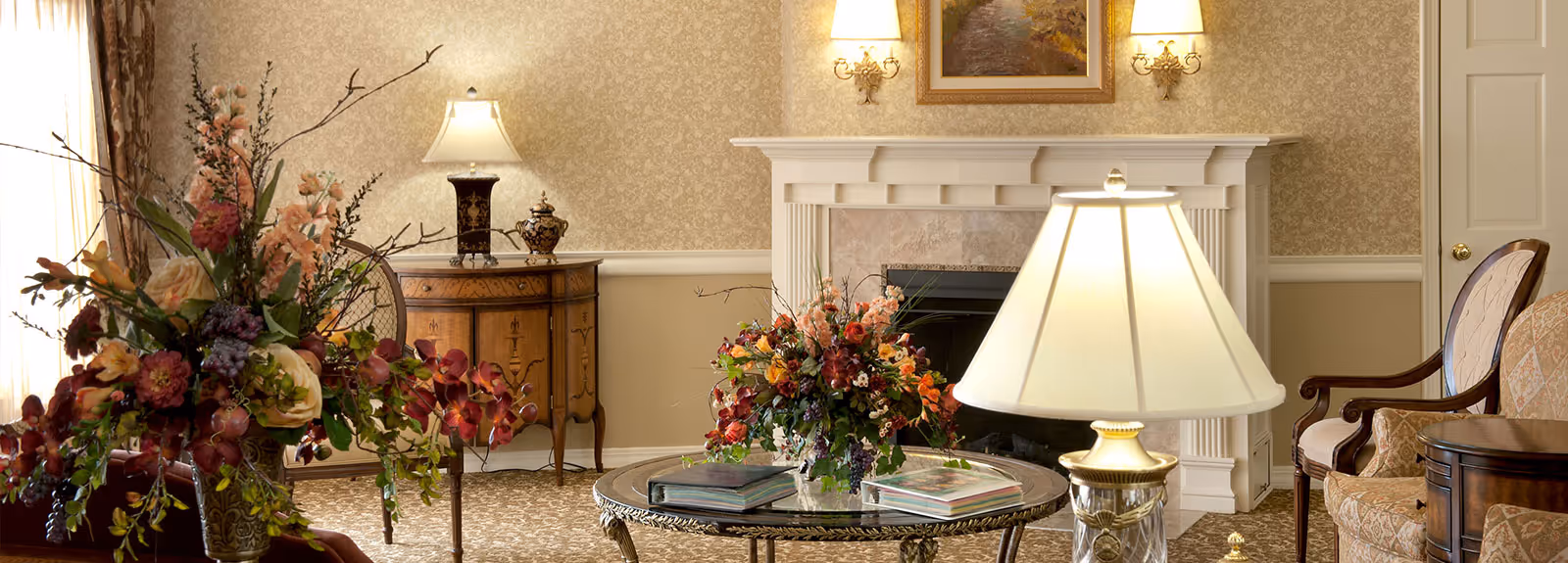 Comfortably furnished living room with a fireplace, floral arrangements, lamps, and seating around a glass coffee table.