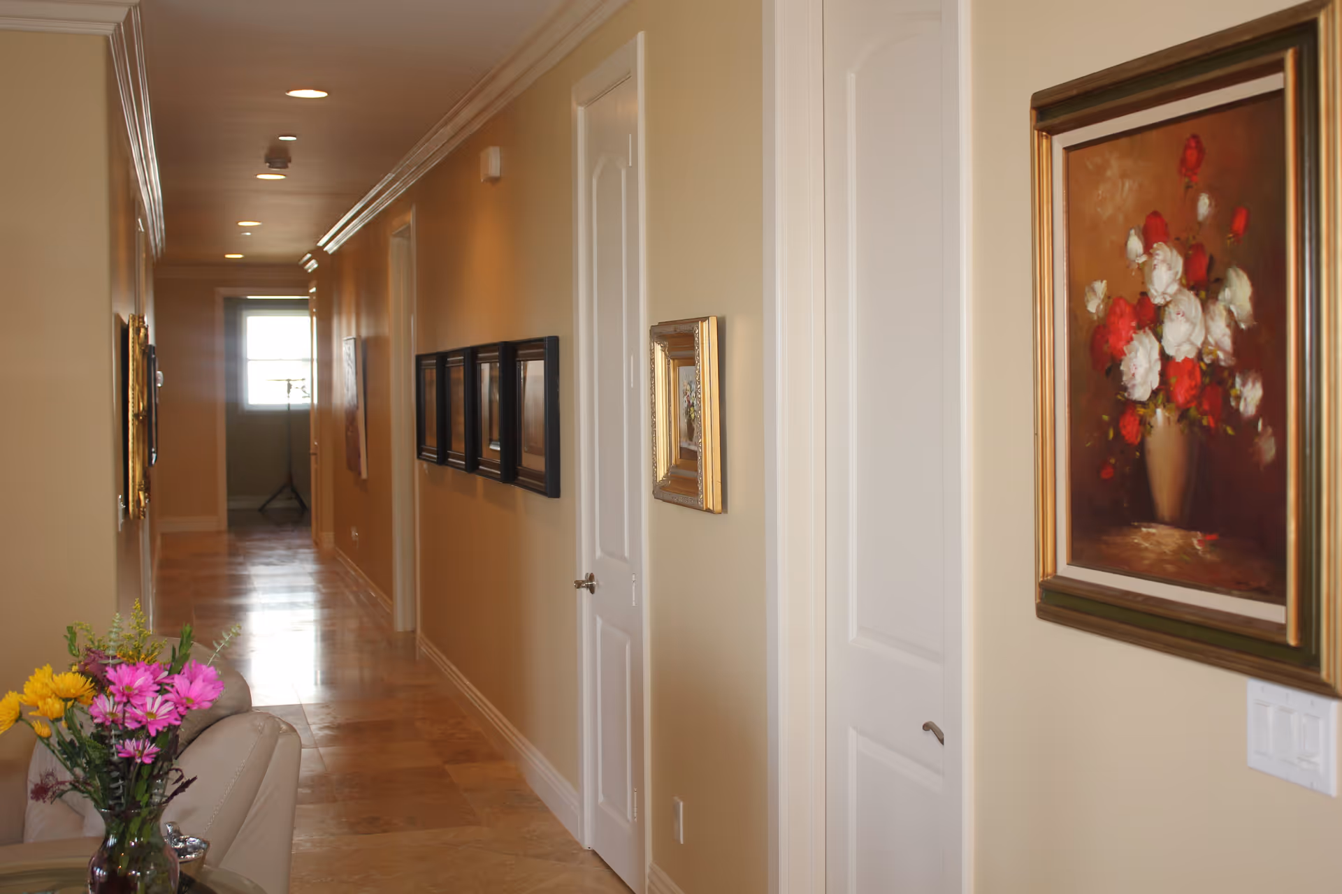 Well-lit interior hallway with framed artwork on the walls, several doors, tiled floor, and a small floral arrangement by a couch.