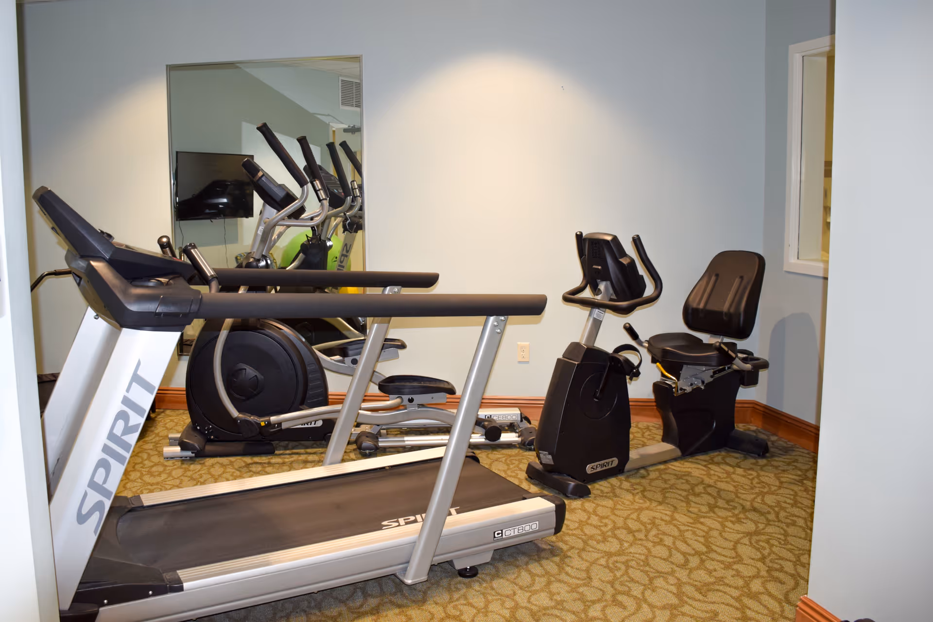 A small exercise room with a treadmill, an elliptical machine, and a recumbent stationary bike. The room has light-colored walls, a patterned carpet, a large mirror on one wall, and a window on another wall.