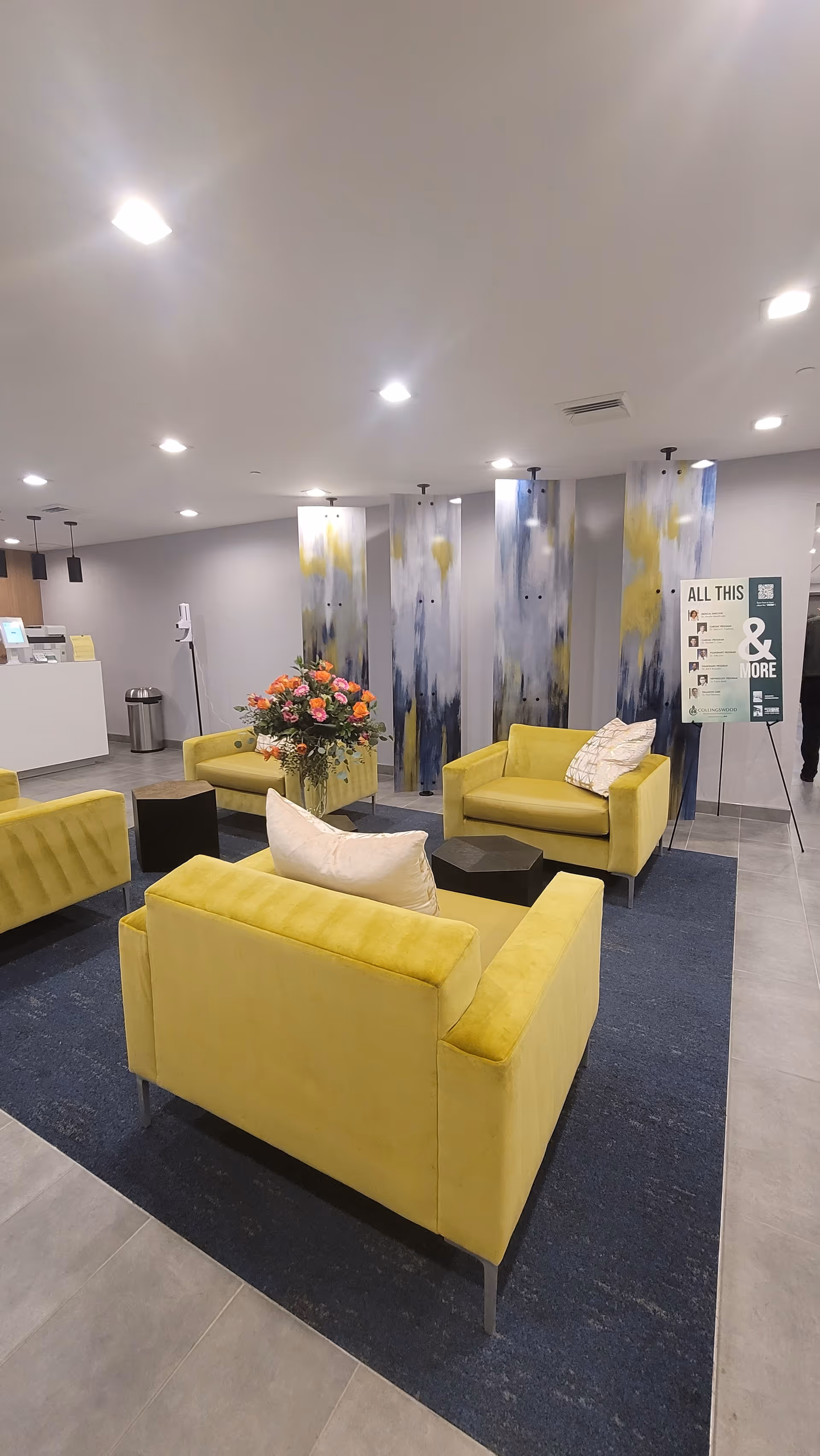 A modern indoor seating area with four yellow armchairs arranged around two small black tables on a dark blue rug. There is a bouquet of flowers on one of the tables. The background features abstract vertical panels with blue, white, and yellow patterns, a white reception desk, a trash can, and a signboard with text.