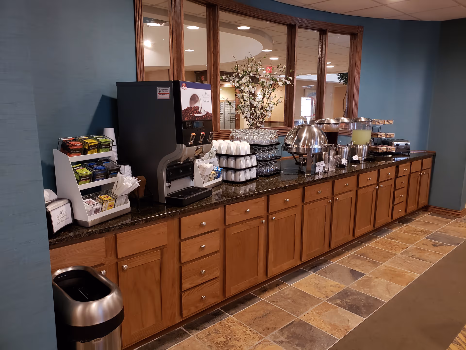 A long wooden counter with multiple cabinets underneath, featuring a coffee and tea station with a beverage dispenser, stacks of cups, tea bags, a juice dispenser, covered food trays, and a decorative flower arrangement in front of large windows reflecting the interior space.
