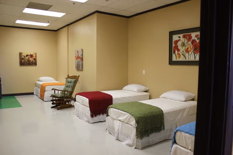 A room with four single beds lined up against a beige wall, each bed has a white sheet and a colorful blanket at the foot. There is a cushioned rocking chair between two of the beds. The walls are decorated with framed floral paintings. The floor is light-colored and the ceiling has fluorescent lights.