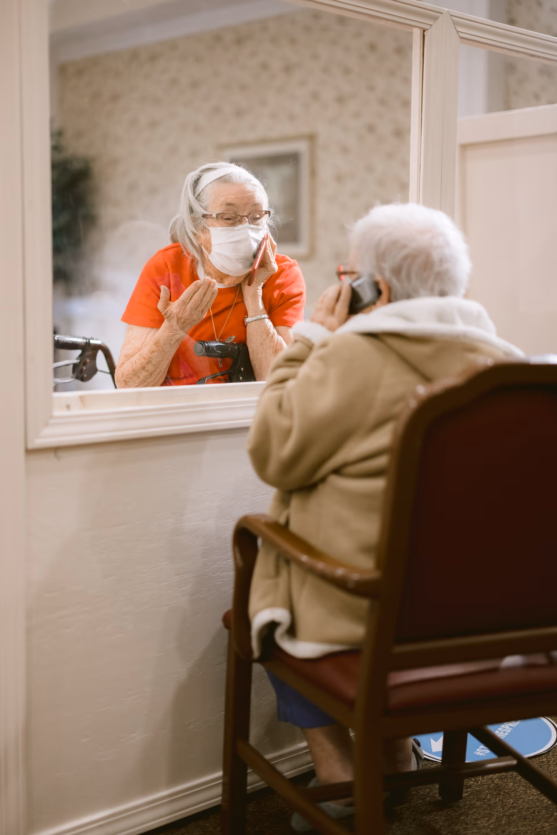 An elderly woman wearing a face mask and red shirt is seen through a glass partition, talking on a phone. On the other side of the glass, another elderly woman wearing a beige coat and glasses is seated on a chair, also talking on a phone. They appear to be communicating through the glass in a senior living facility.