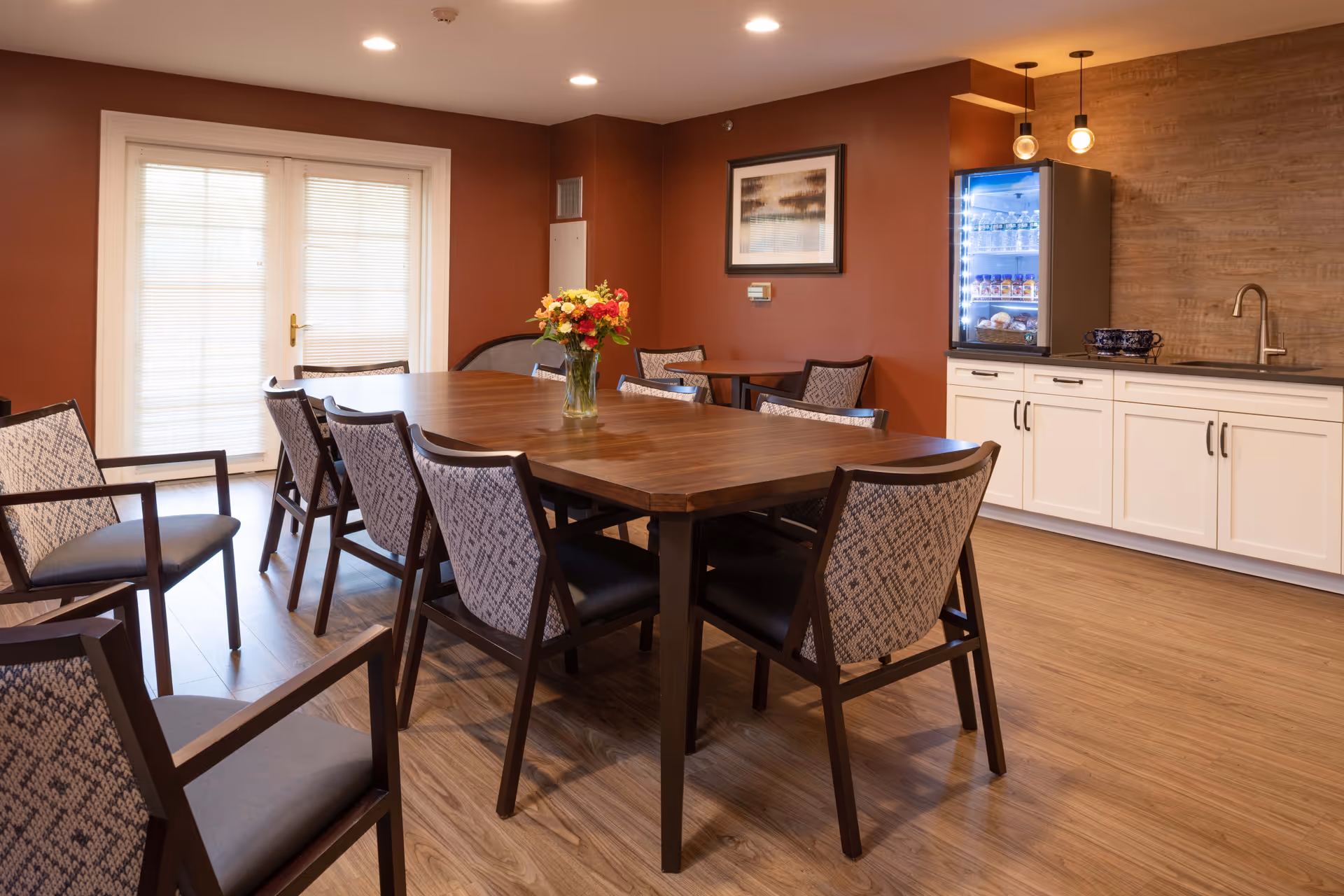 A dining room with a large wooden table surrounded by eight patterned chairs. A vase with colorful flowers is placed in the center of the table. The room has warm brown walls, a wooden floor, and a set of double glass doors with blinds. On one side, there is a countertop with white cabinets, a small sink, and a glass-front refrigerator stocked with beverages. Two pendant lights hang above the countertop.