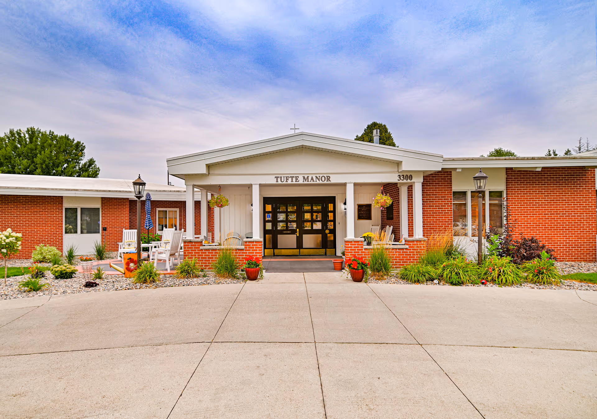Front exterior view of Tufte Manor, a single-story brick building with a white entrance porch featuring columns and hanging flower pots. There are outdoor chairs and tables on the left side, surrounded by landscaped plants and shrubs under a partly cloudy sky.