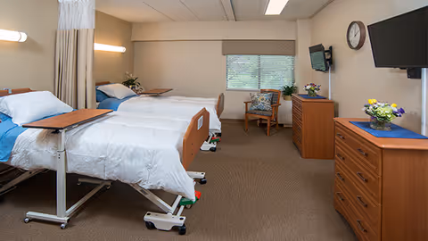 A bright shared retirement facility bedroom with two hospital-style beds, dressers, a wall-mounted TV, a chair, and a window.