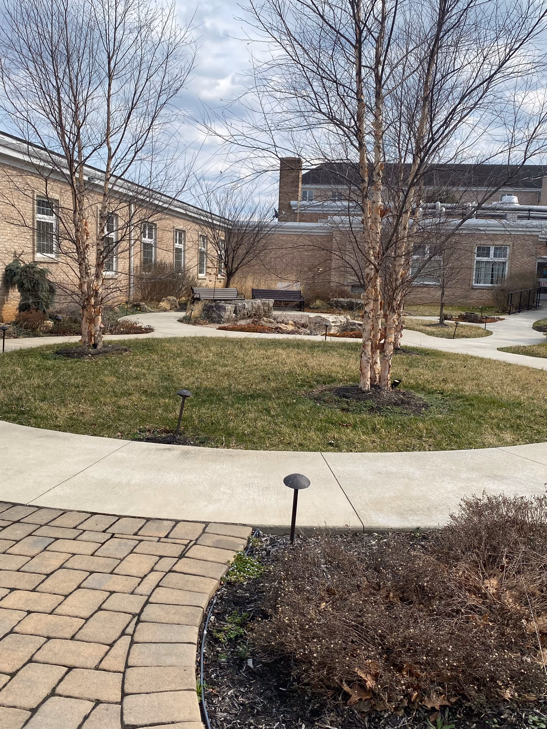 Outdoor courtyard area with paved walkways, grass, leafless trees, and benches in front of a brick building under a cloudy sky.