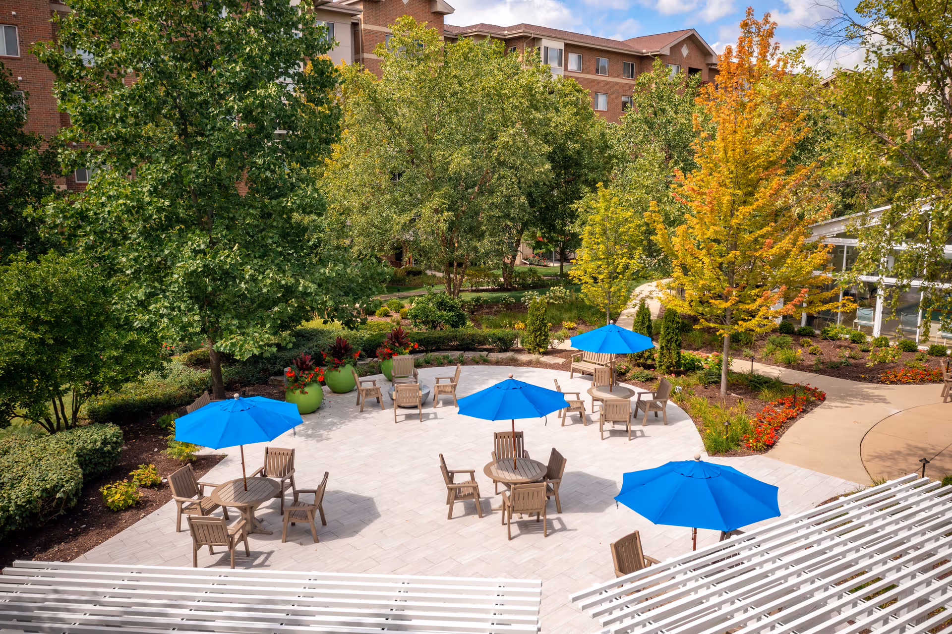 Outdoor patio area with several round tables and wooden chairs, each table shaded by a bright blue umbrella. The patio is surrounded by lush green trees and colorful plants, with a multi-story brick building in the background under a partly cloudy sky.