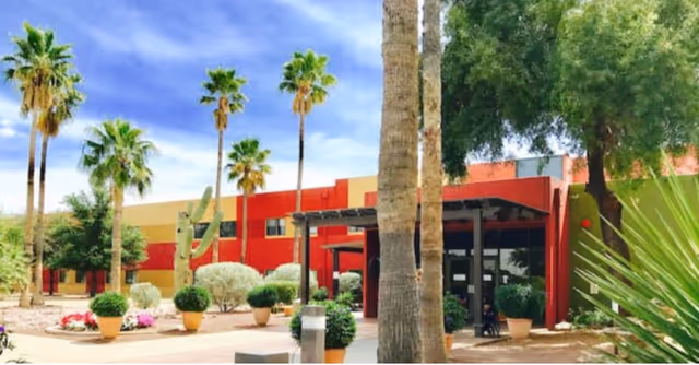 Exterior view of Sandstone of Tucson facility showing a modern building with red and beige walls, surrounded by palm trees, desert plants, and potted shrubs under a partly cloudy blue sky.