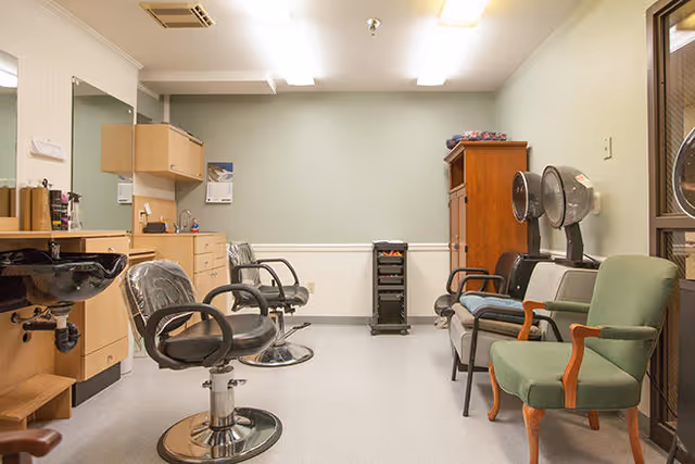 Interior of a salon or hair care room with two black salon chairs in front of a counter with a sink and mirror. On the right side, there are two hair drying chairs with hooded dryers and two green upholstered chairs. The walls are light green with white wainscoting, and there is a wooden cabinet in the back corner.