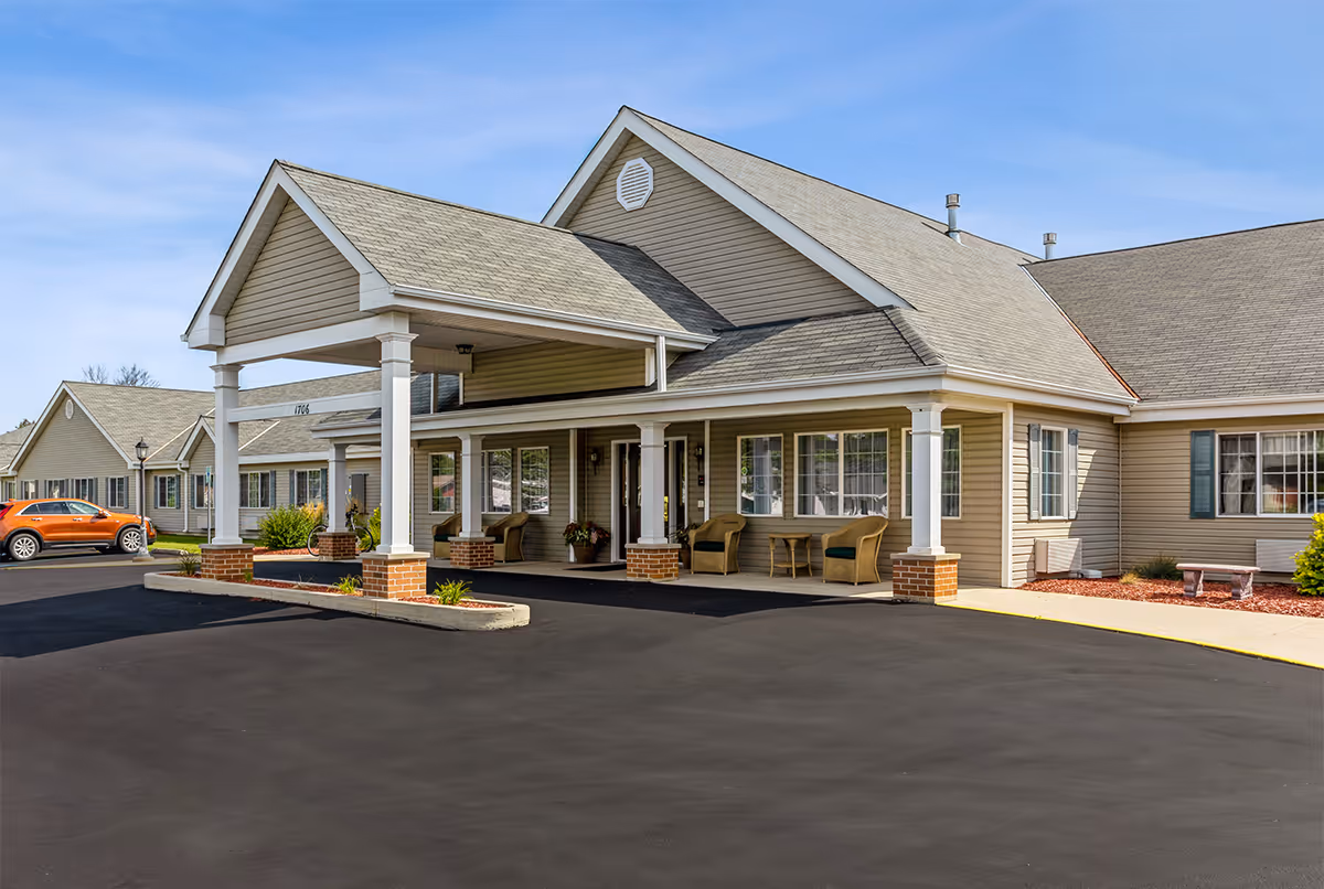 Exterior view of LakeHouse New Holstein facility showing a beige building with a covered entrance supported by white columns, outdoor seating with chairs and a table, a paved driveway, and a parked orange car under a clear blue sky.