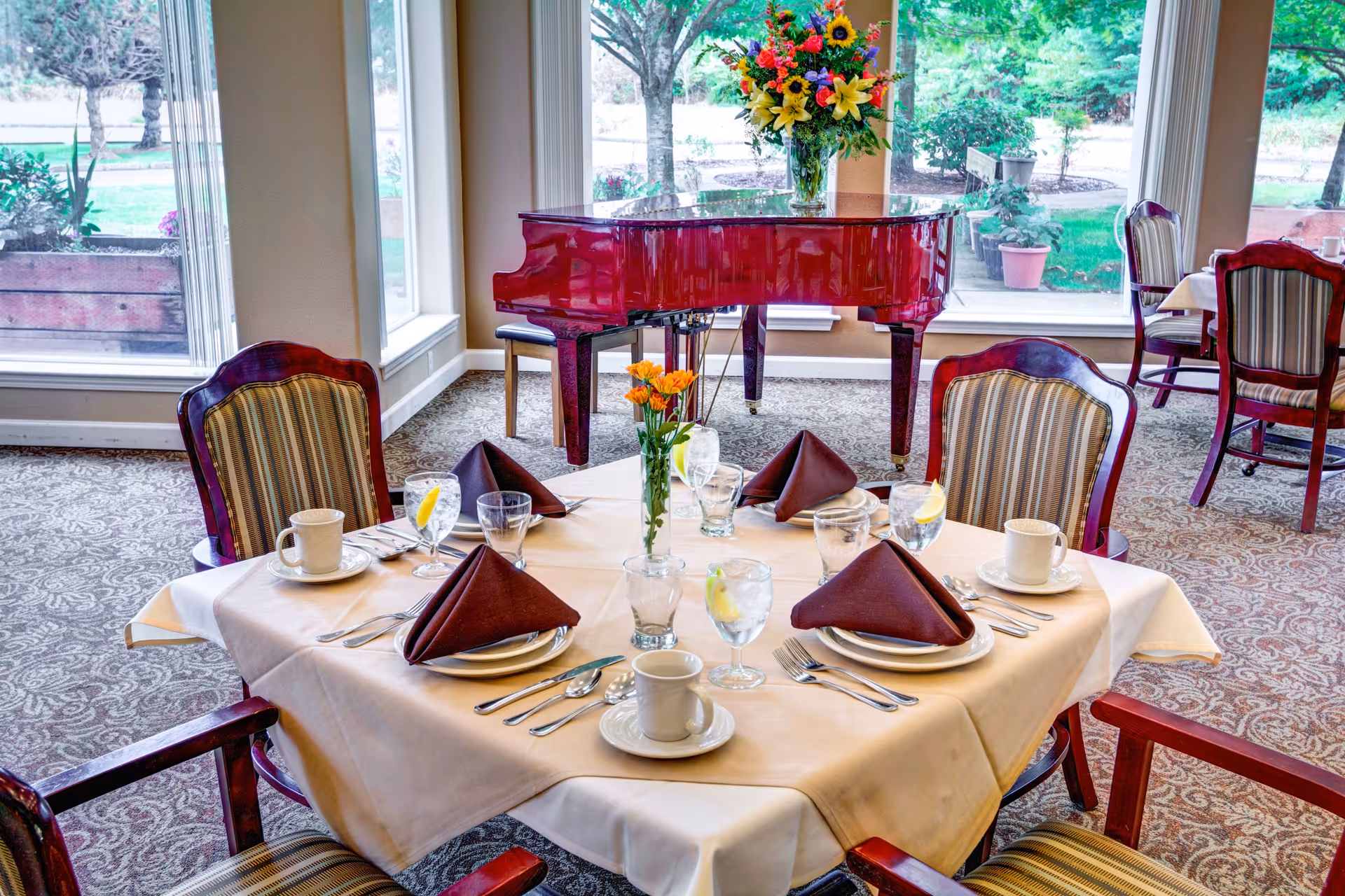 A dining table set for four with white tablecloth, brown folded napkins, cups, glasses with lemon slices, and silverware. Behind the table is a red grand piano with a colorful flower arrangement on top. Large windows show a green outdoor garden area.