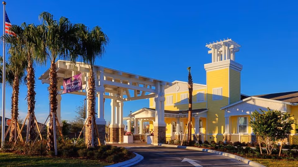 Front entrance of a yellow senior living building with a covered porte-cochere, palm trees, and a clear blue sky.