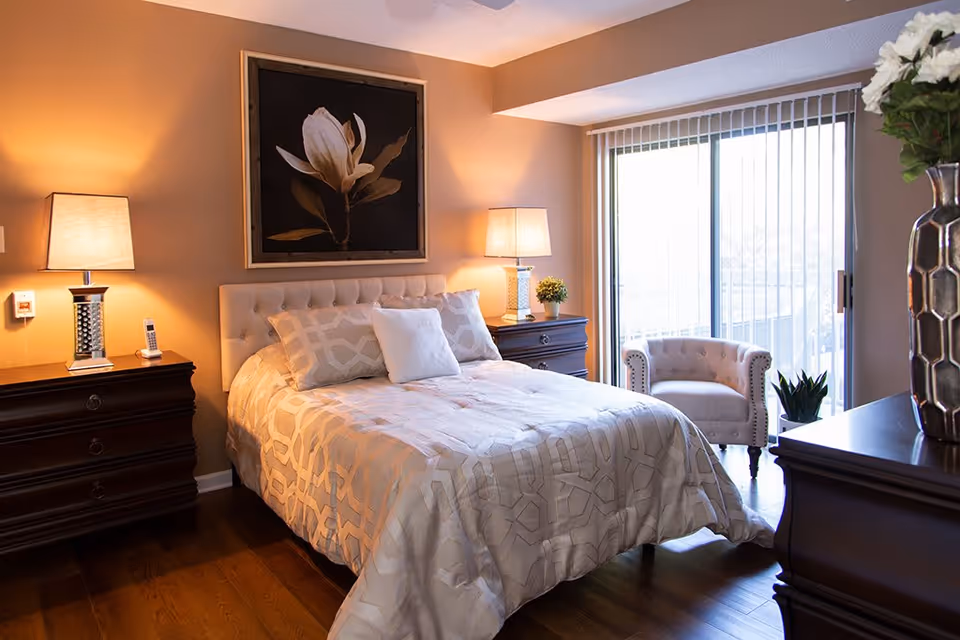A cozy bedroom with a neatly made bed featuring a beige patterned comforter and pillows. There are two dark wooden nightstands on either side of the bed, each with a lamp. Above the bed hangs a large framed painting of a white flower. To the right, there is a sliding glass door with vertical blinds leading to a balcony. A beige upholstered armchair is placed near the door, and a dark wooden dresser with a vase of flowers is visible in the foreground.
