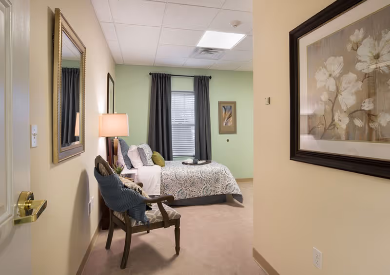 A furnished bedroom with a patterned bed, wooden chair, bedside lamp, framed artwork, and a window with dark curtains.