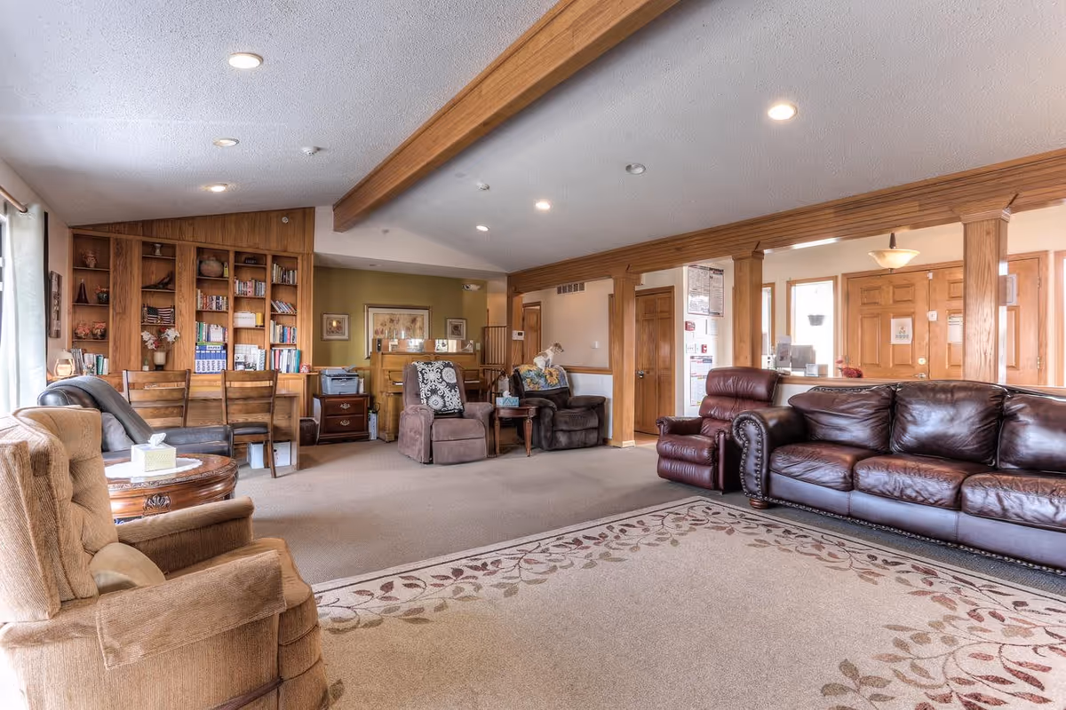 A spacious senior living facility common area with comfortable seating including a brown leather sofa, recliners, and armchairs. The room features a large patterned area rug, wood-paneled walls, built-in bookshelves filled with books and decorative items, and a desk with chairs. The ceiling has recessed lighting and wooden beams, and there are windows with curtains allowing natural light to enter.