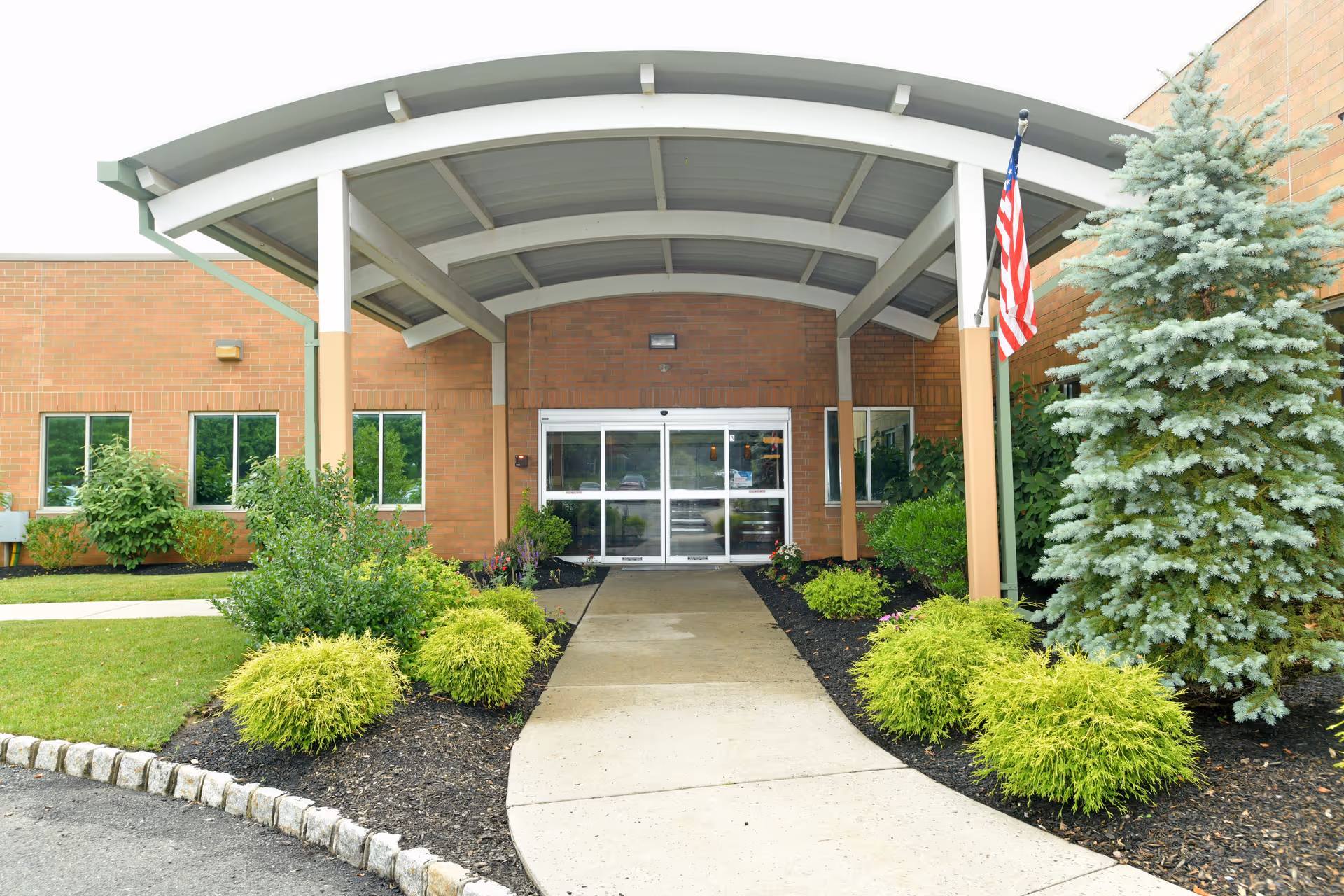 Entrance to a brick building with a covered walkway supported by white beams. There are landscaped bushes and a small tree on either side of the concrete path leading to automatic glass doors. An American flag is mounted on the right side near the entrance.