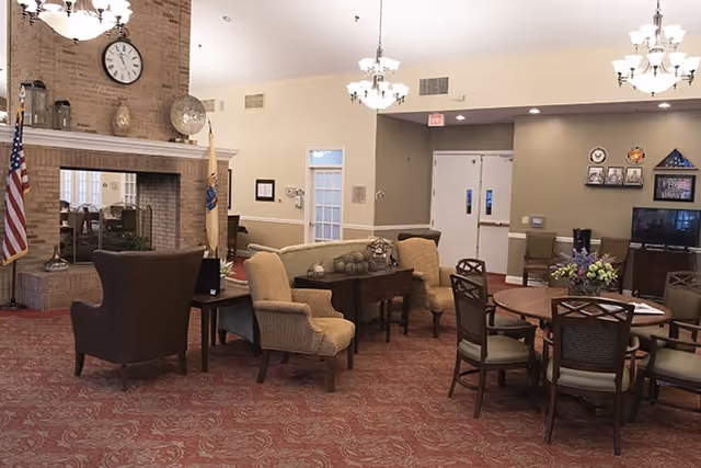 Spacious senior living common room with clustered seating, round dining tables, a brick fireplace, and chandeliers.