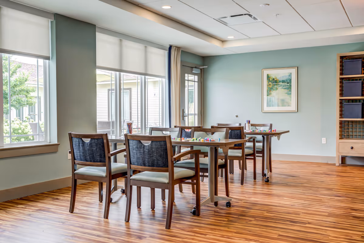 Bright activity/dining room with wooden tables and chairs, large windows, and a framed painting on the wall.
