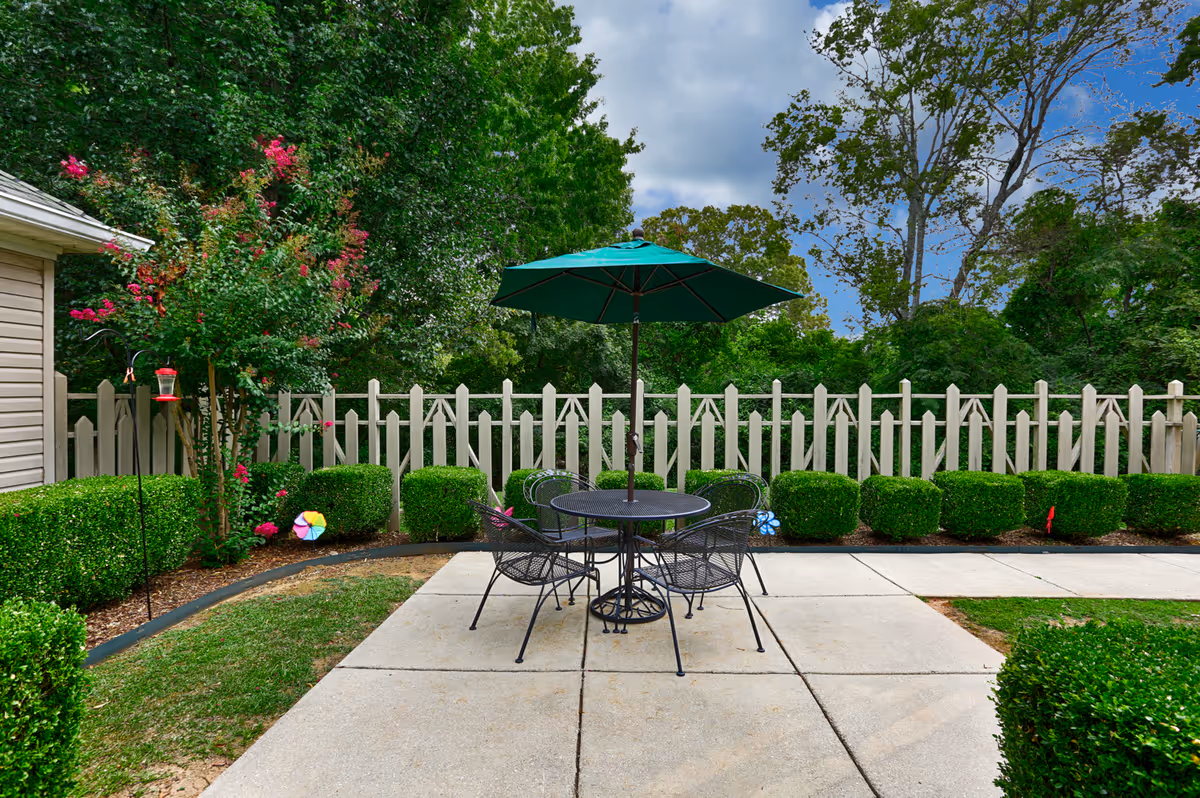 Outdoor patio area with a round metal table and four matching chairs under a green umbrella. The patio is surrounded by neatly trimmed bushes, a white picket fence, and trees with green foliage. There is a small garden area with flowering plants and a bird feeder on the left side.