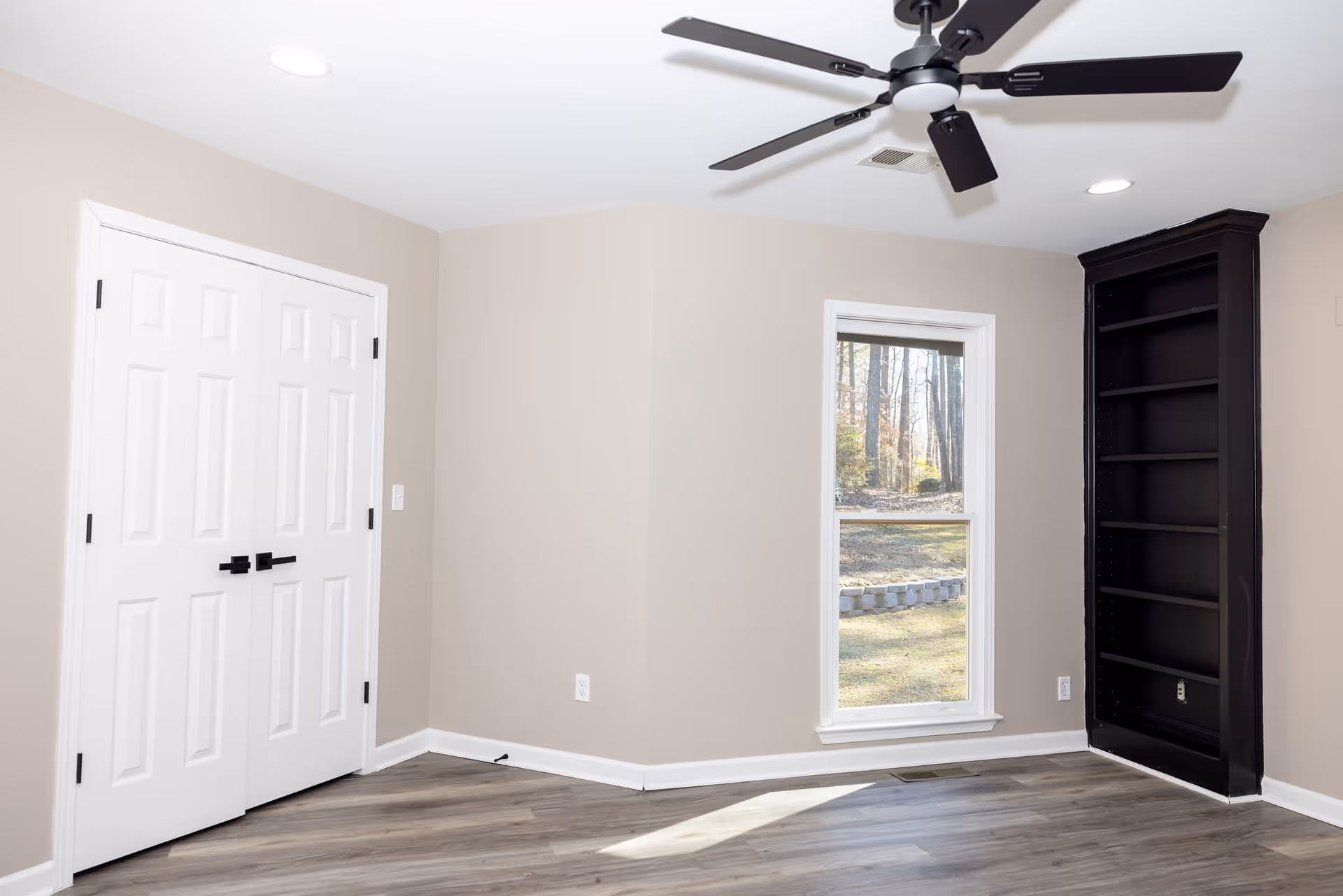 Empty room with beige walls, a white double-door closet, a large window showing an outdoor view with trees and grass, a black built-in bookshelf, a ceiling fan with black blades, and wood-style flooring.