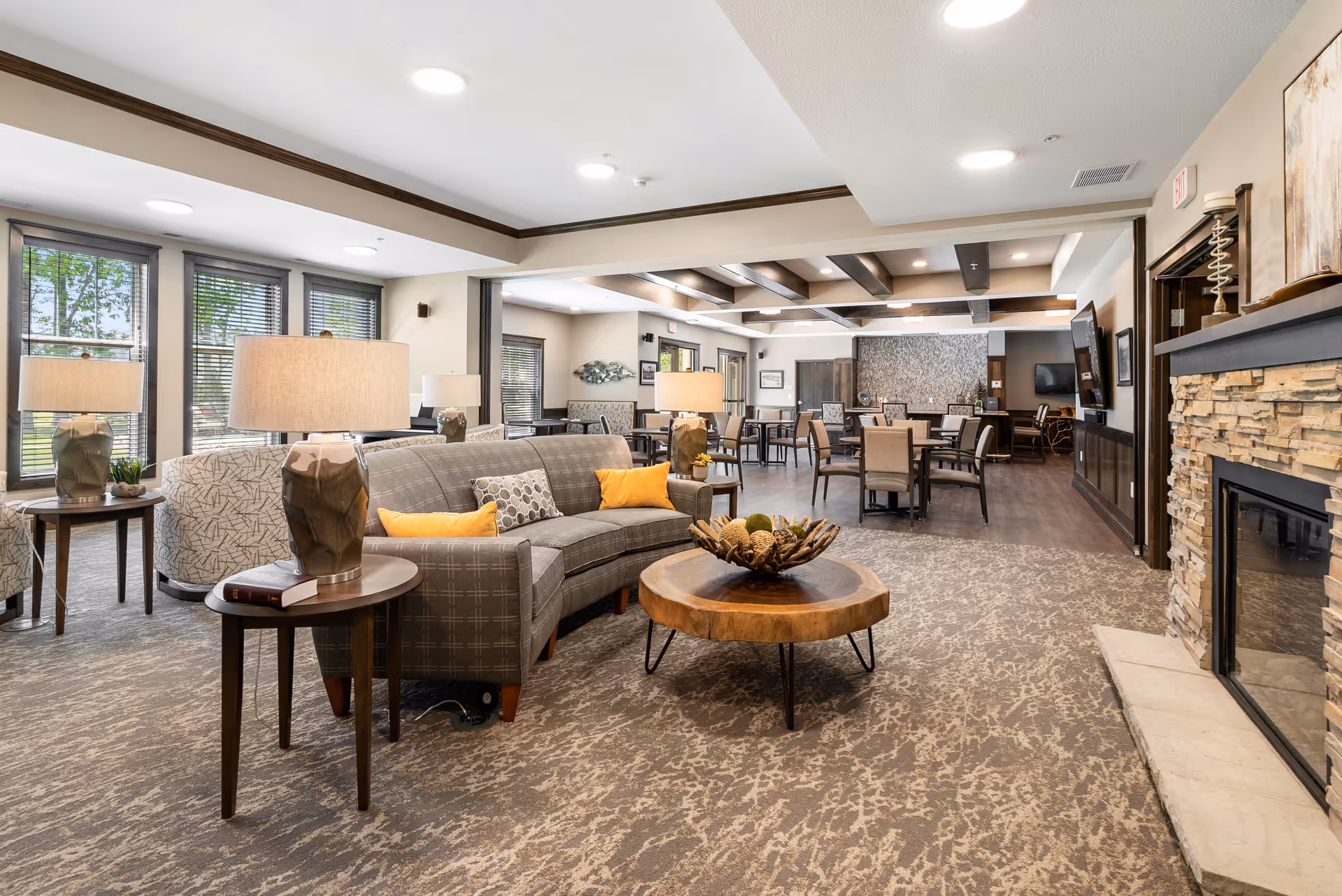 Spacious senior living common area with sofas, side tables and lamps, a coffee table, fireplace, and a dining area in the background.