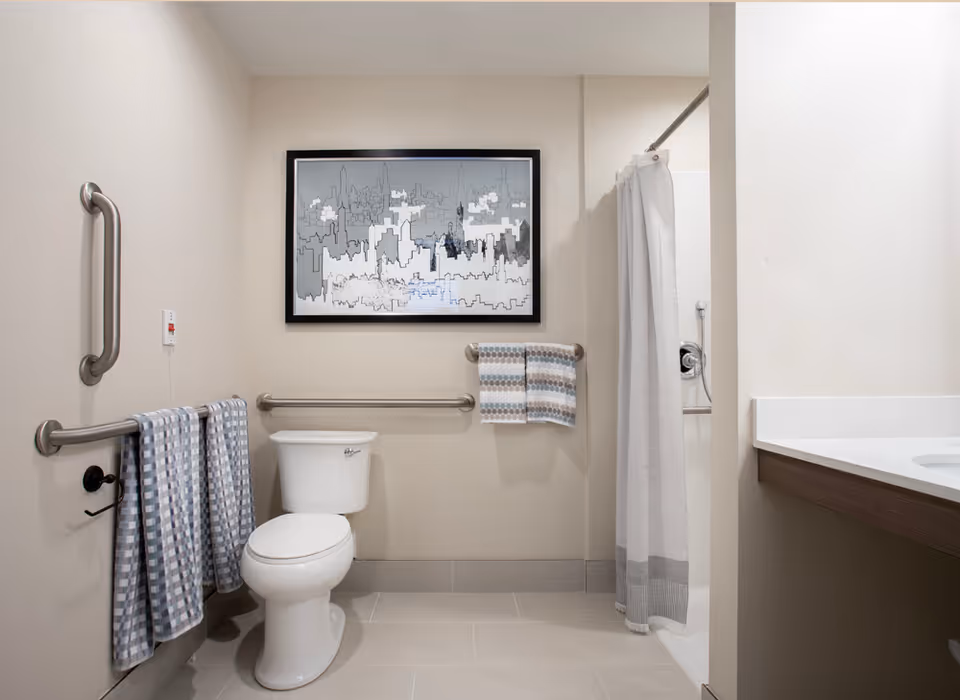 A clean and accessible bathroom featuring a white toilet with grab bars on the walls around it, a shower with a white curtain, a countertop with a sink on the right, and two sets of patterned towels hanging on the grab bars. A framed abstract cityscape artwork is mounted on the wall above the toilet.