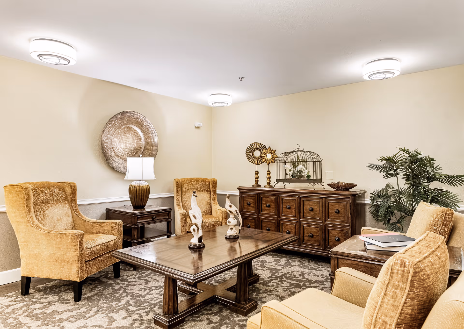 A cozy living room area with four mustard-yellow upholstered armchairs arranged around a wooden coffee table. The coffee table has two decorative sculptures on it. Behind the chairs, there is a wooden sideboard with multiple drawers, decorated with ornamental items including a birdcage and two sunburst sculptures. A table lamp is placed on a small side table between two chairs. The room has beige walls, a patterned carpet, and ceiling lights. There is also a green potted plant in the corner.