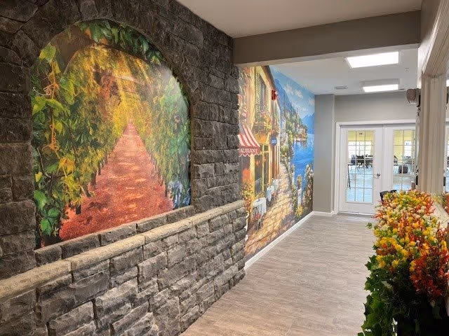 Interior hallway with stone-textured wall featuring a mural of a leafy archway and a scenic waterfront village. The opposite wall has a colorful flower arrangement and a door leading to a bright room with tables and chairs.