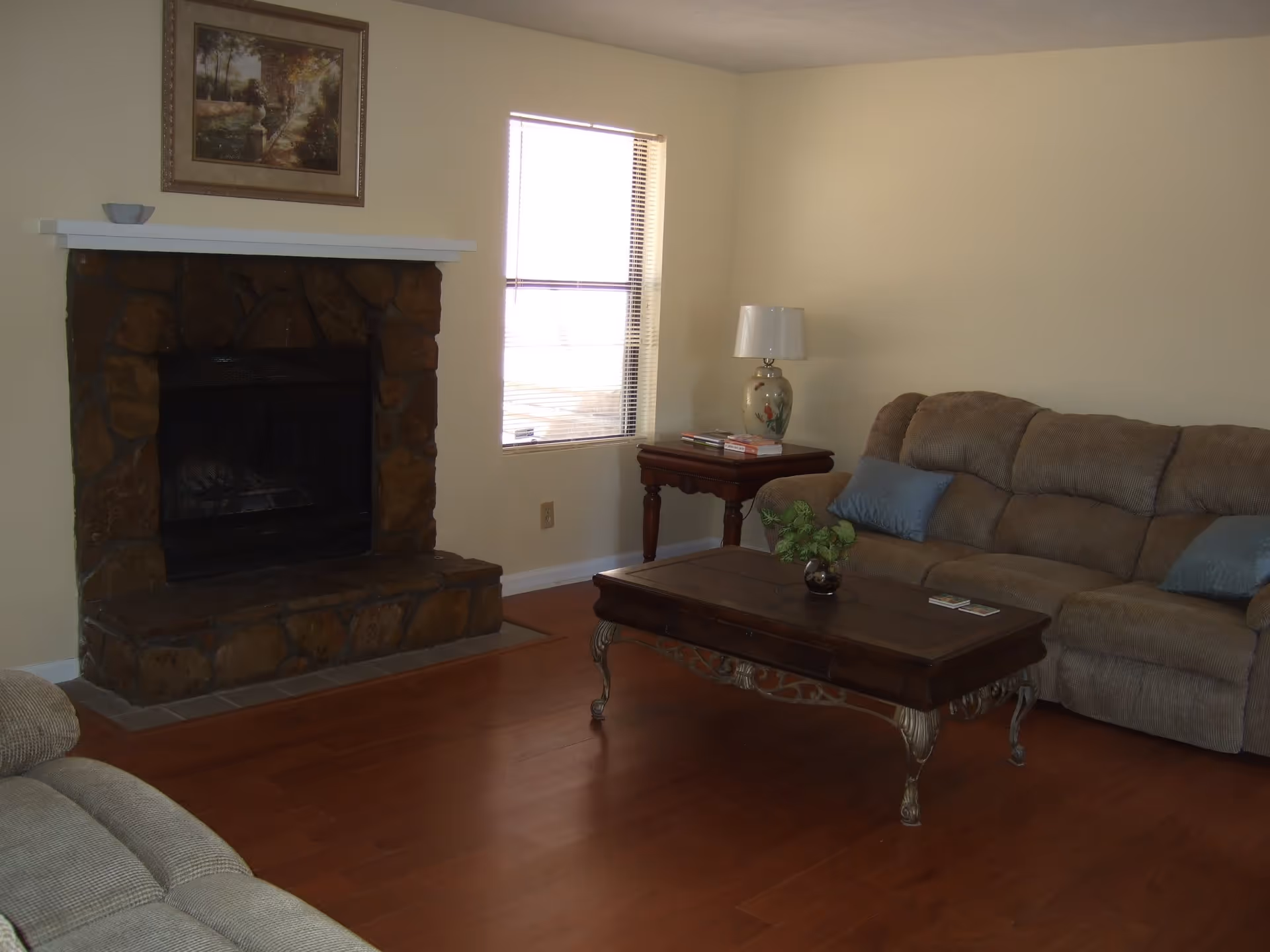 A living room with a stone fireplace, a beige upholstered sofa, wooden coffee table and side table beneath a window.
