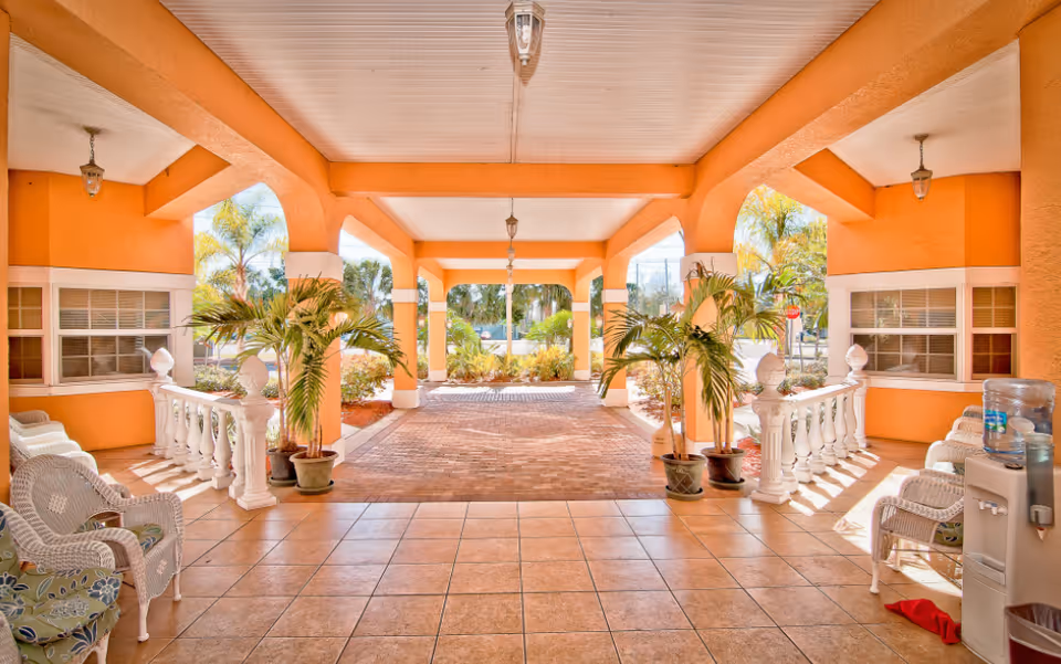 Covered outdoor seating area with orange walls and white ceiling, featuring wicker chairs with cushions, potted palm plants, and a water dispenser on the right side. The area opens to a brick-paved driveway with greenery and palm trees visible in the background.