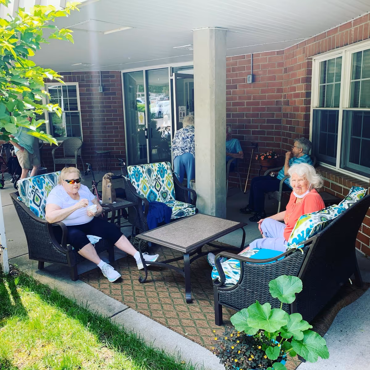 Several elderly individuals sitting and relaxing on cushioned outdoor patio furniture under a covered area with brick walls and large windows. Some are seated on patterned chairs and sofas around a central table, while others are in the background near the entrance door. Green plants and grass are visible in the foreground.
