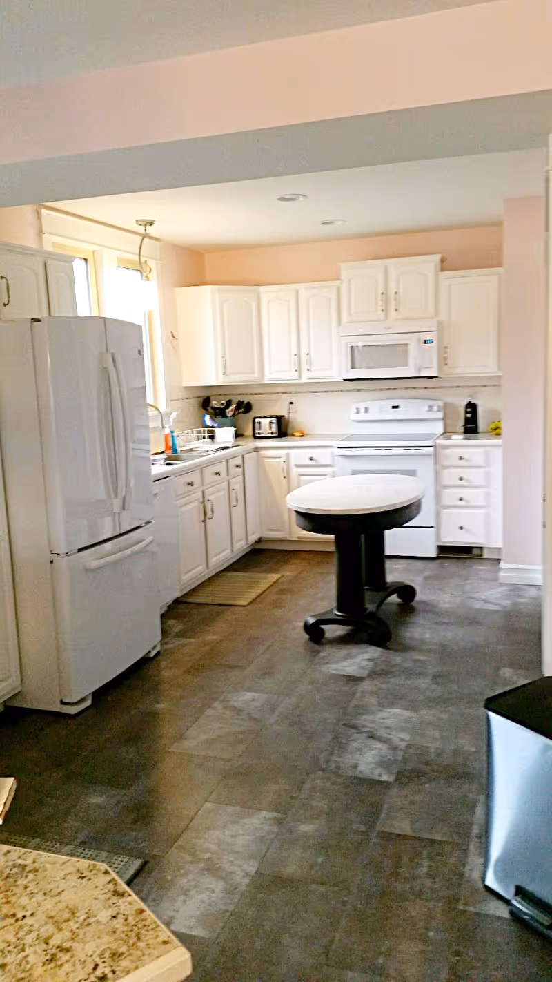 A clean kitchen with white cabinets, a white refrigerator, a white stove with a microwave above it, a small round table on wheels in the center, and various kitchen utensils on the countertop near a window.