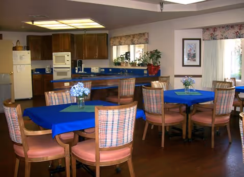 Interior view of a dining area with several tables covered in blue tablecloths and decorated with small flower arrangements. The room has wooden chairs with plaid upholstery, a kitchen area with wooden cabinets, a refrigerator, microwave, and coffee maker. There are windows with floral valances and framed artwork on the walls.