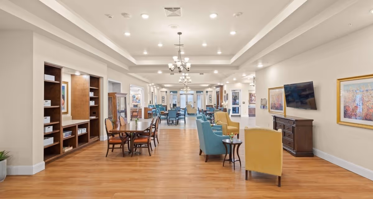 A spacious, well-lit common area in a senior living facility with wooden flooring, multiple seating arrangements including blue and yellow armchairs, wooden tables with chairs, a wall-mounted TV, framed artwork on the walls, and built-in shelving units on the left side.
