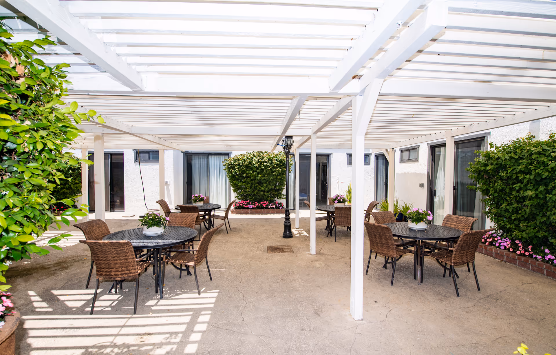 Outdoor patio area with a white pergola overhead, several round tables with wicker chairs, potted plants on the tables, and greenery and flowers along the edges. The patio is adjacent to a building with sliding glass doors and windows.