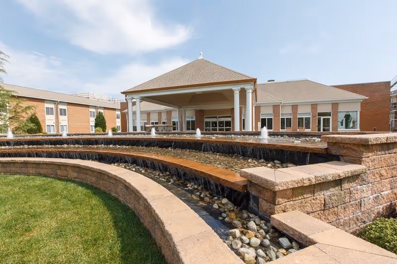 Outdoor view of Oak Bridge Terrace At Manor House featuring a tiered stone water fountain with flowing water and small jets, surrounded by green grass and a large building with a covered entrance supported by white columns under a partly cloudy sky.