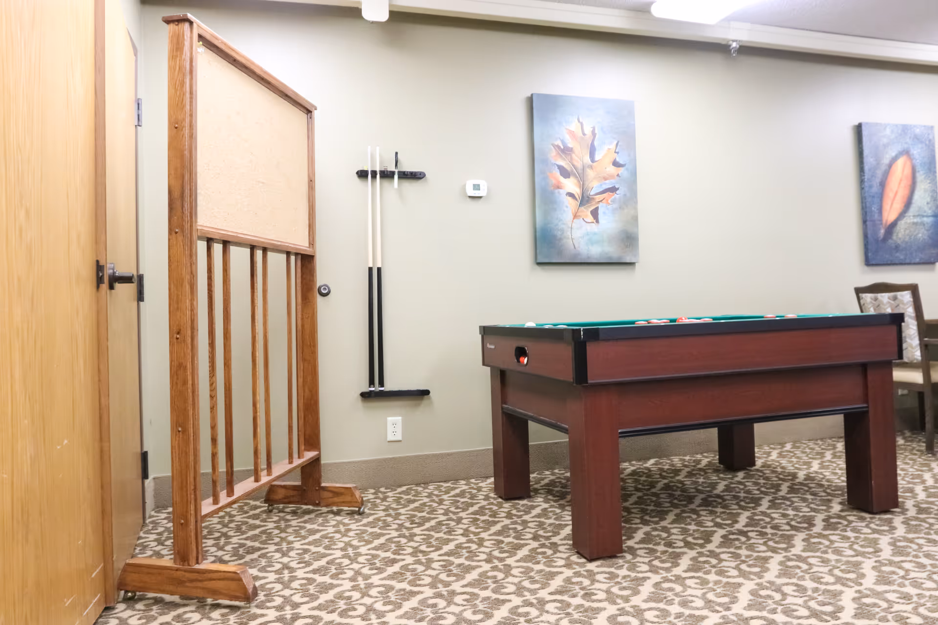 A game room with a pool table, two pool cues mounted on the wall, a wooden divider, patterned carpet, and two framed leaf paintings on the wall.