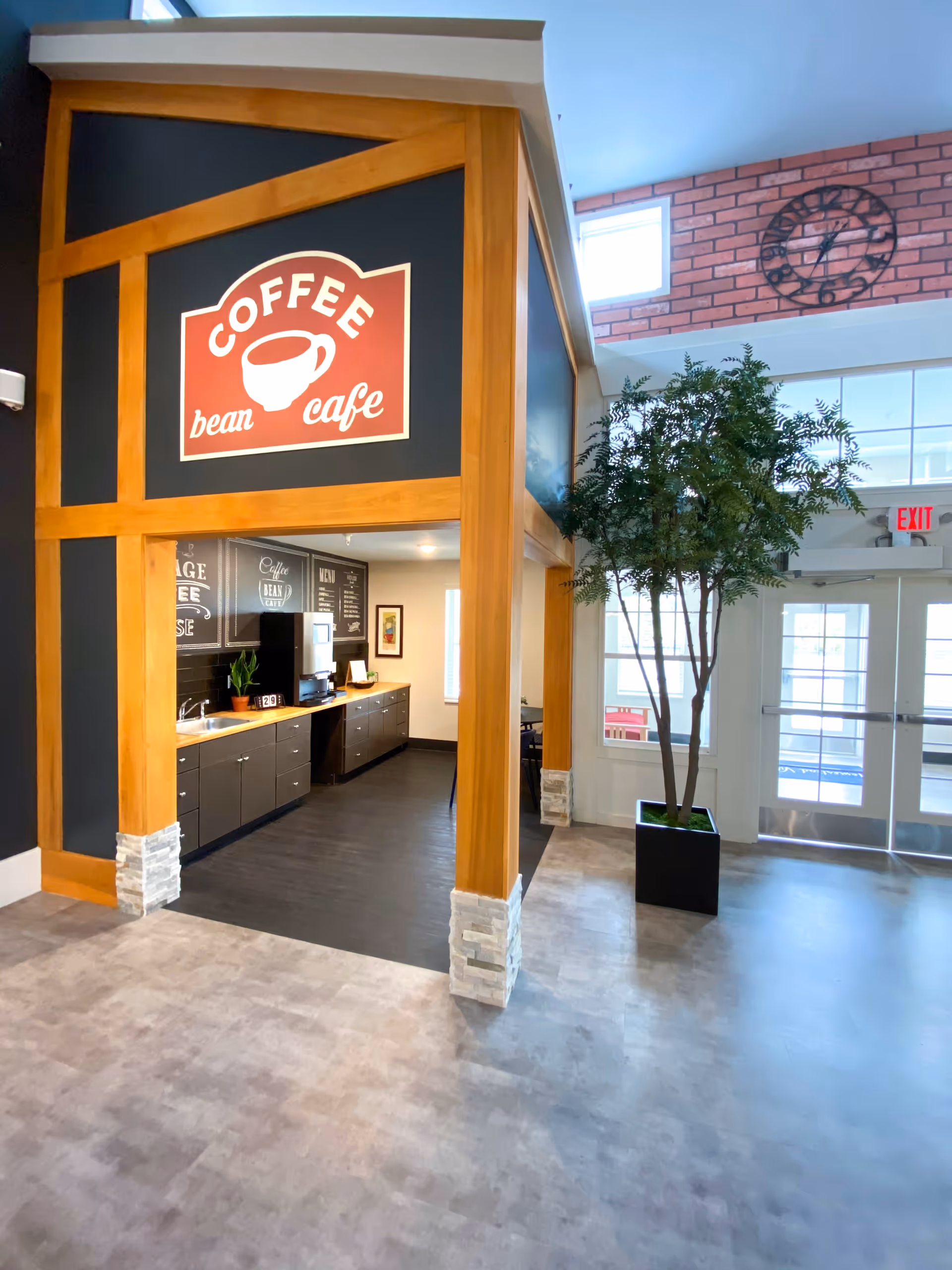 Interior view of a coffee cafe area inside a facility, featuring a wooden framed structure with a sign that reads 'Coffee bean cafe'. Inside the cafe area, there is a counter with cabinets, a coffee machine, and some plants. To the right, there is a large potted tree, windows, and a door with an exit sign above it. A large clock is mounted on a brick wall above the door.