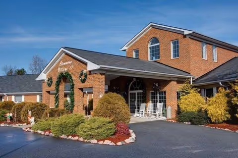 Brick senior living center with arched entrance and rocking chairs near landscaped shrubs.